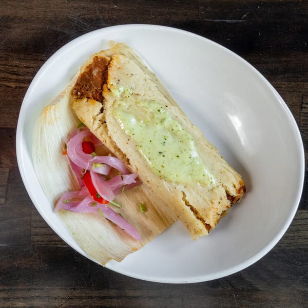 Tamale topped with green sauce and pickled red onions, served on a corn husk.