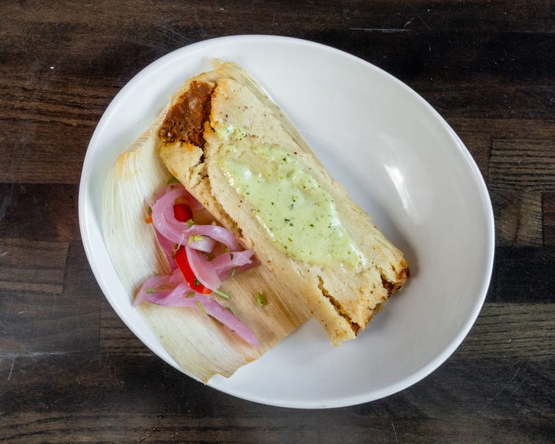 Tamale topped with green sauce and pickled red onions, served on a corn husk.