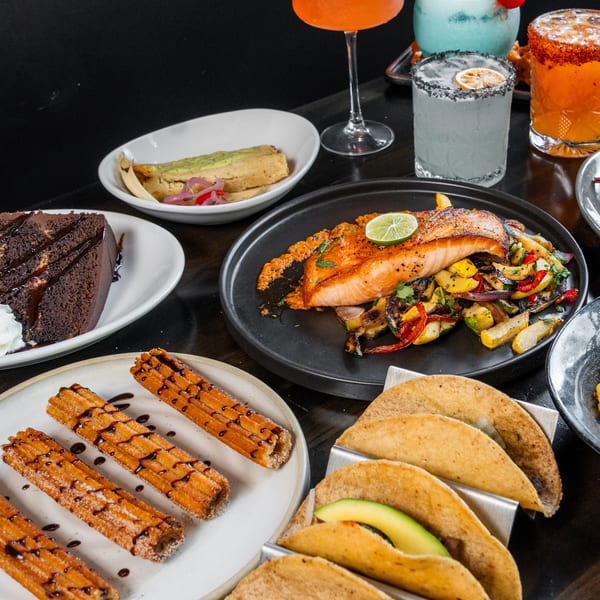 Table filled with various Mexican dishes like tacos, salmon, churros, cake, and colorful cocktails.