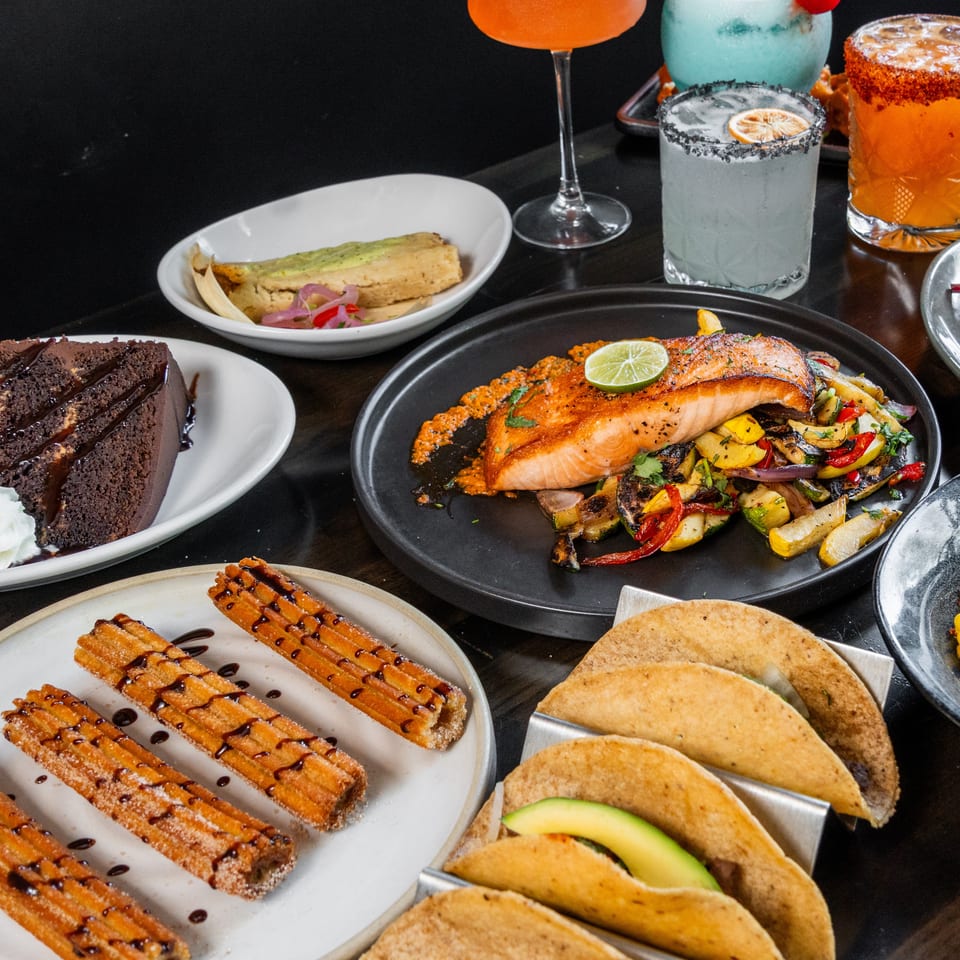 Table filled with various Mexican dishes like tacos, salmon, churros, cake, and colorful cocktails.