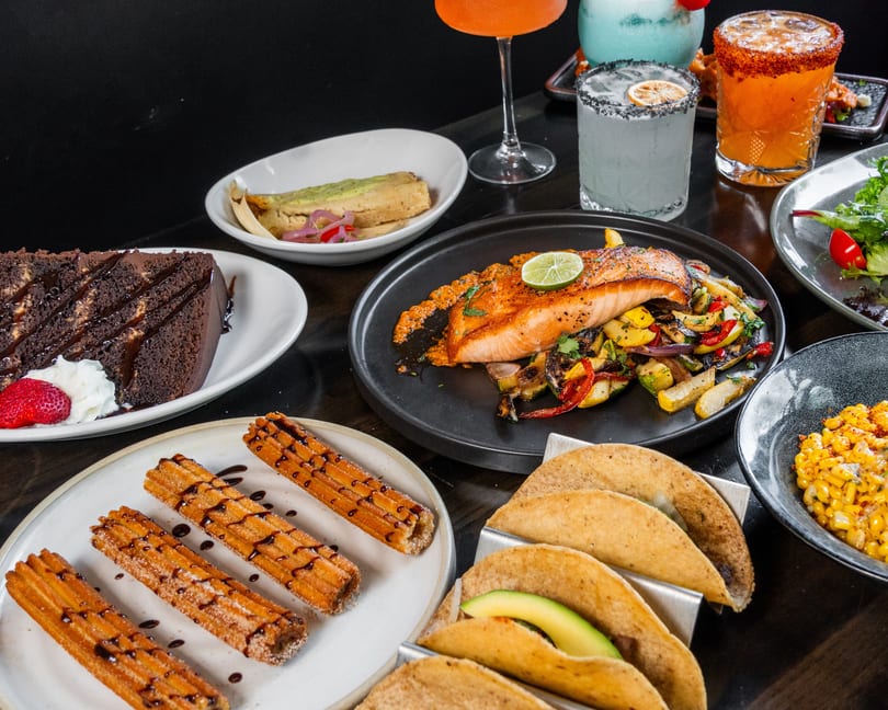 Table filled with various dishes including salmon, tacos, churros, chocolate cake, and colorful cocktails.