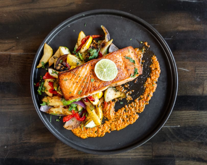 Pan-seared salmon with roasted vegetables, a lime slice, and orange sauce on a black plate.