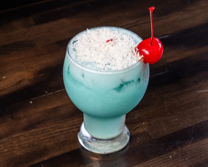 Blue cocktail with shredded coconut and a red cherry on a dark wooden table.