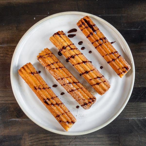 Four golden churros, drizzled with chocolate and sprinkled with sugar, on a white plate.