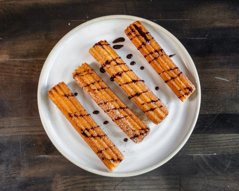 Four golden churros, drizzled with chocolate and sprinkled with sugar, on a white plate.