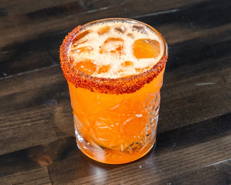 Vibrant orange cocktail with a chili-salt rim and ice in a cut glass.