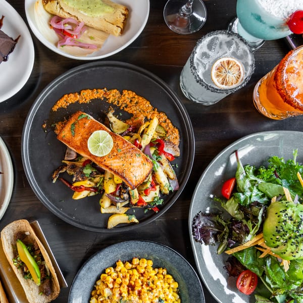 A vibrant spread of salmon, tacos, salad, desserts, and cocktails on a dark wooden table.