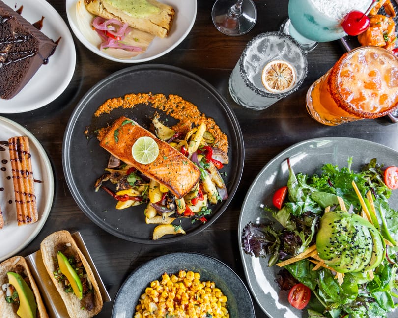 A vibrant spread of salmon, tacos, salad, corn, churros, cake, tamales, and colorful margaritas on a table.