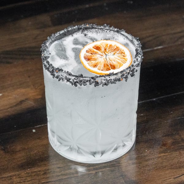 A clear cocktail in a textured glass with a black rim and dried orange slice.