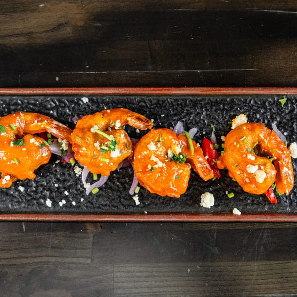 Four orange-glazed shrimp with crumbled cheese, chives, and red onion on a black rectangular plate.