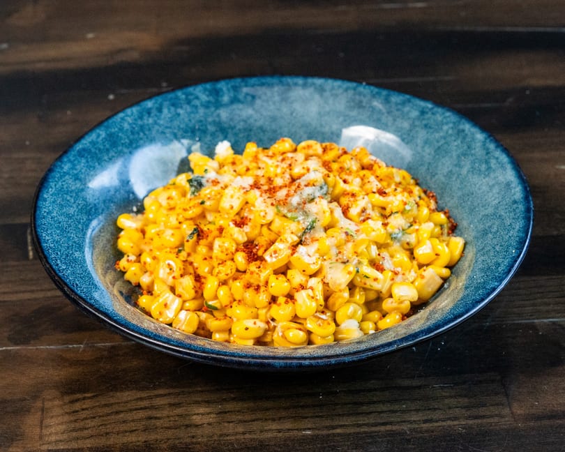 A blue bowl filled with seasoned corn, topped with cheese and red chili flakes.