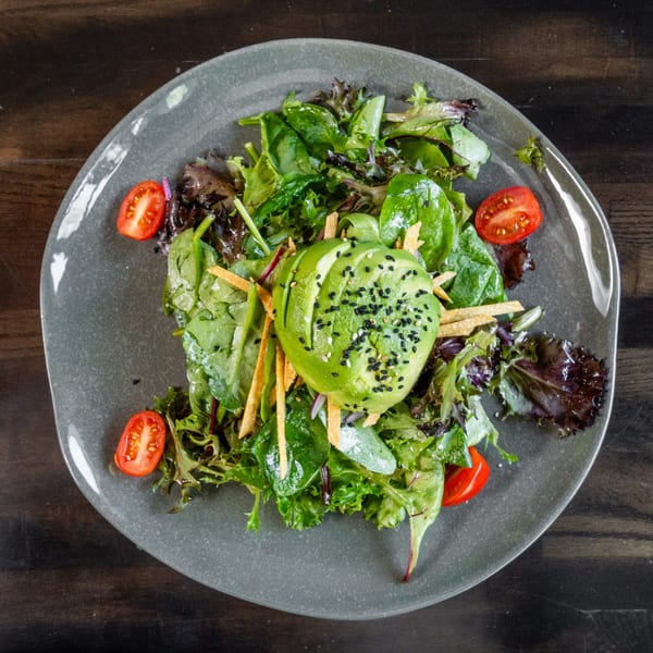 Mixed green salad with sliced avocado, sesame seeds, cherry tomatoes, and tortilla strips on a gray plate.