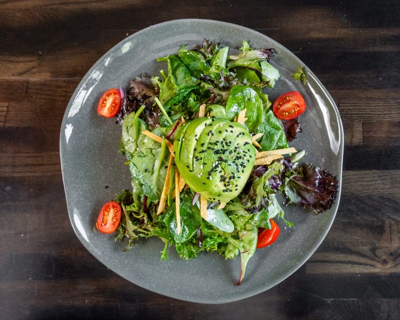 Mixed green salad with sliced avocado, sesame seeds, cherry tomatoes, and tortilla strips on a grey plate.