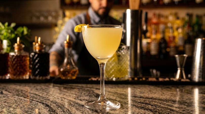 A pale yellow cocktail with a lemon twist on a marble bar, with a bartender in the background.