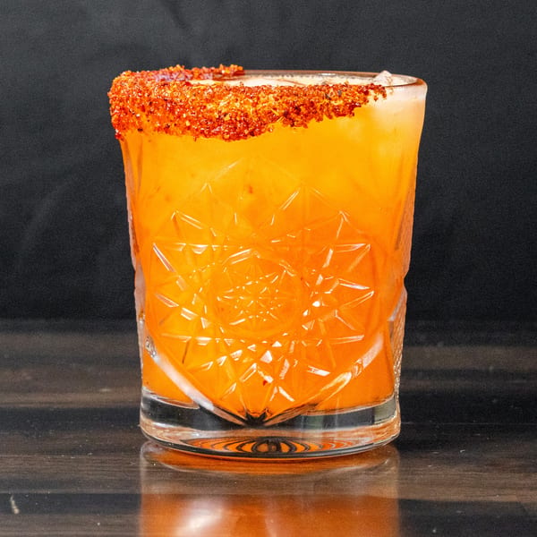 Bright orange cocktail in a faceted glass, rimmed with red chili flakes.