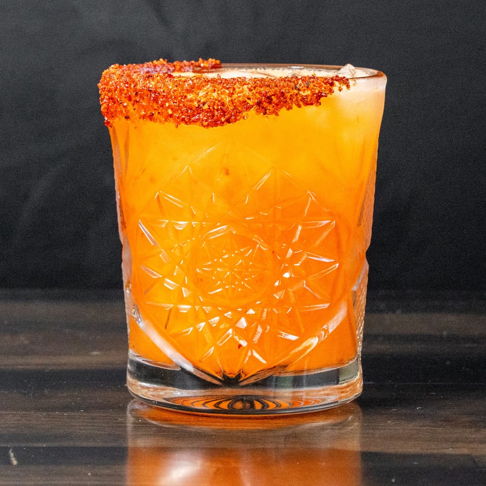 Bright orange cocktail in a faceted glass, rimmed with red chili flakes.