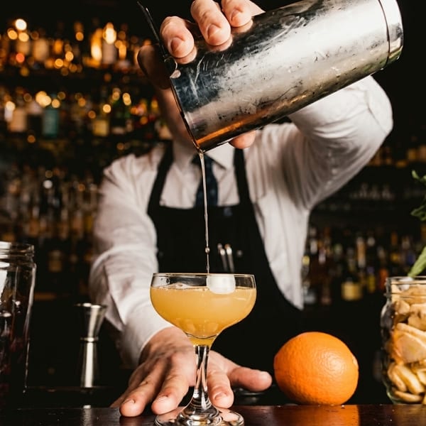 Bartender pouring a cocktail from a shaker into a glass, with fresh herbs and fruit on the bar.