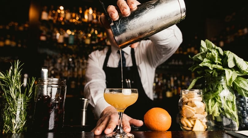 Bartender pouring a cocktail from a shaker into a glass, with fresh herbs and fruit on the bar.