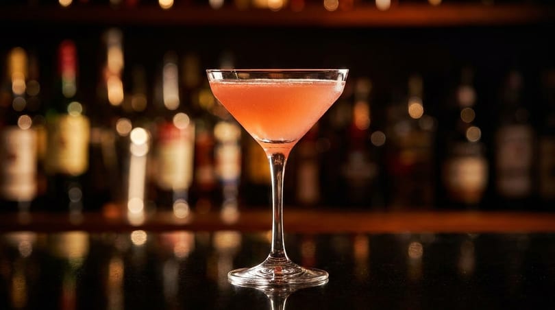 A pink cocktail in a martini glass sits on a dark bar with blurred liquor bottles in the background.