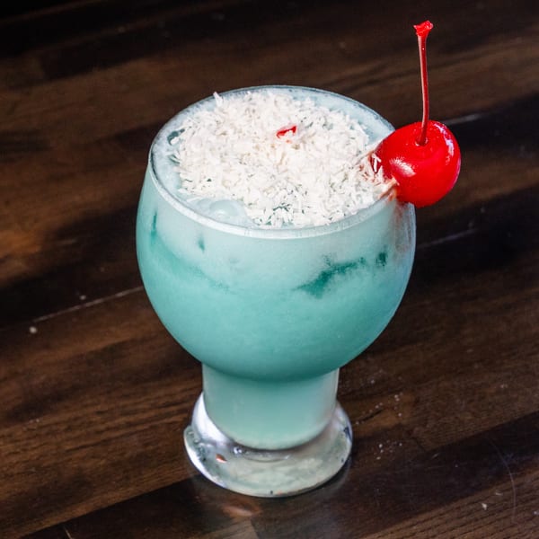 Blue cocktail with shredded coconut and a bright red cherry on a dark wood surface.