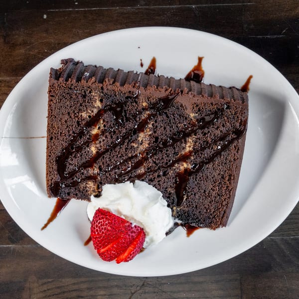 Slice of chocolate cake with chocolate drizzle, whipped cream, and a strawberry on a white plate.