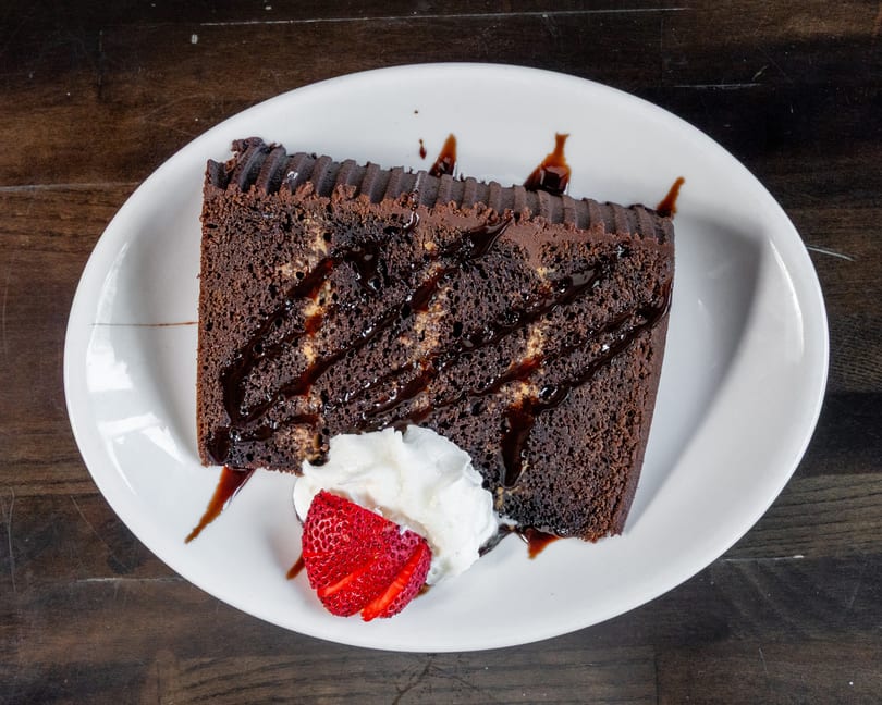 Slice of chocolate cake with chocolate drizzle, whipped cream, and a strawberry on a white plate.
