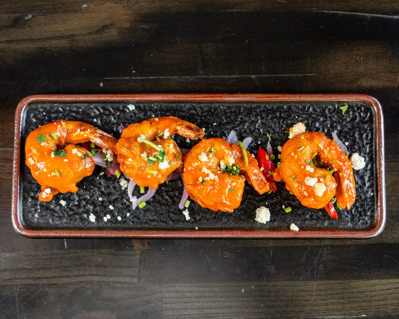 Four orange shrimp with sauce, herbs, cheese, red onion, and peppers on a black rectangular plate.