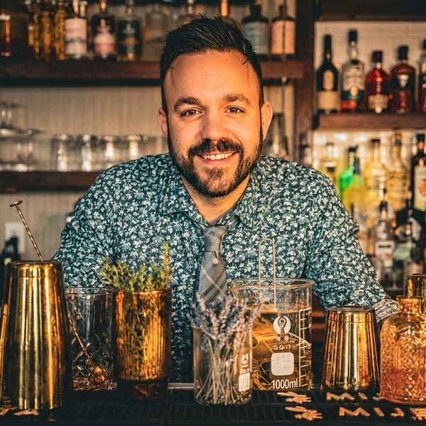 Smiling bartender in a patterned shirt and tie, standing behind a bar with bottles and cocktail tools.