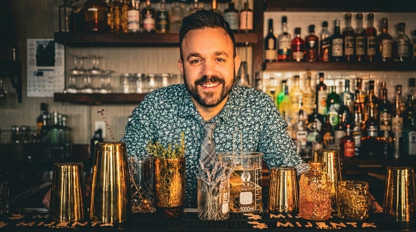 Smiling bartender in a patterned shirt and tie, standing behind a bar with bottles and cocktail tools.