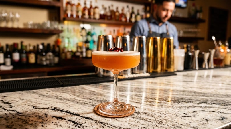 A bartender prepares a cocktail on a granite bar, with bottles and shakers in the background.