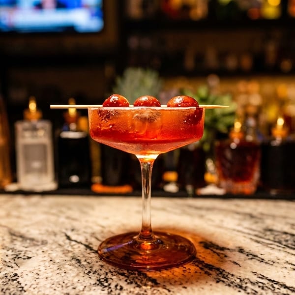 A red cocktail with three cherries on a skewer sits on a marble bar top.