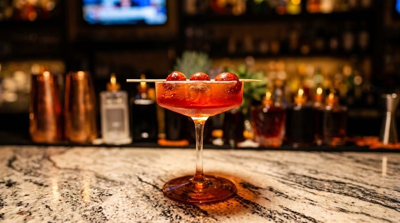 A red cocktail with three cherries on a skewer sits on a marble bar top.
